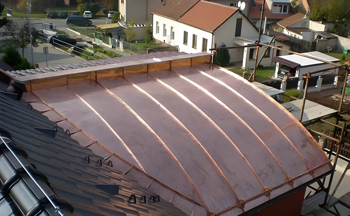 Oblouková falcovaná plechová střecha v měděném odstínu, pohled přes hřeben sousední taškové střechy